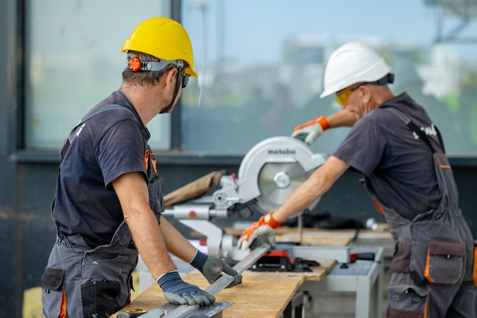 Imagem de dois trabalhadores usando equipamentos de proteção enquanto operam uma serra circular em um ambiente de construção ou fabricação, destacando segurança e colaboração na indústria.