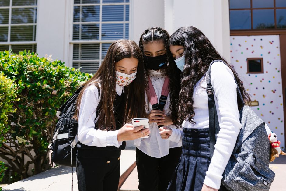 Grupo de estudantes adolescentes usando máscaras e olhando juntos para um celular, ao ar livre, no ambiente escolar.