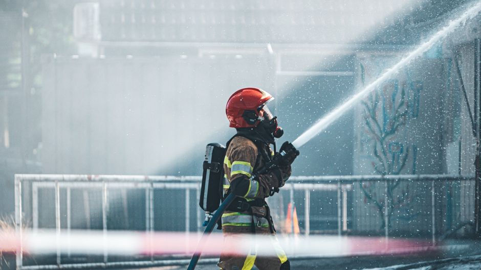 Bombeiro usando equipamento de proteção enquanto combate incêndio com mangueira em ambiente urbano.
