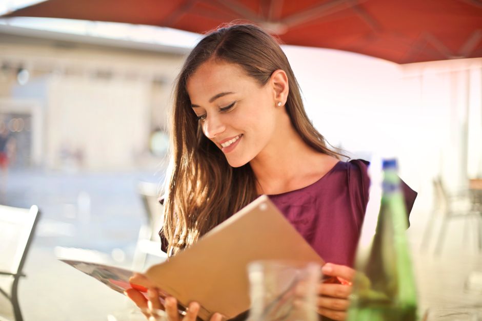 Mulher sorridente lendo um menu em um ambiente ao ar livre, transmitindo descontração e apreciação de momentos de lazer.