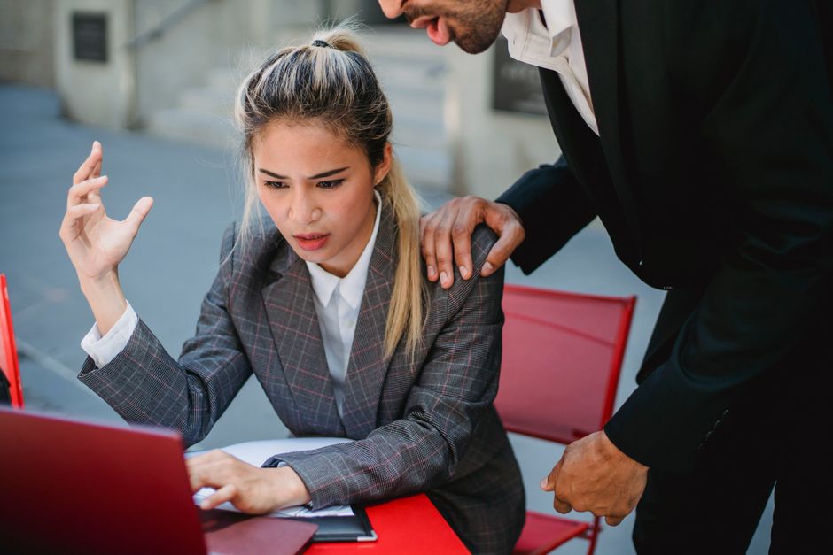 Mulher frustrada diante de seu computador enquanto um colega tenta ajudá-la, demonstrando tensão no ambiente de trabalho.