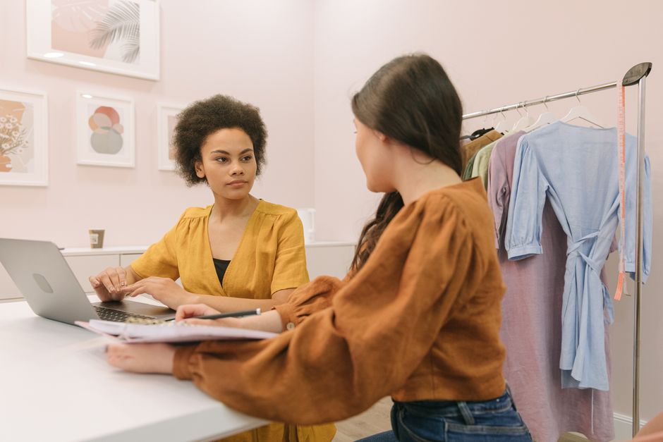 Duas mulheres conversando em um ambiente de roupas, possivelmente em uma loja de moda ou boutique, com uma distinção entre negócios e moda.
