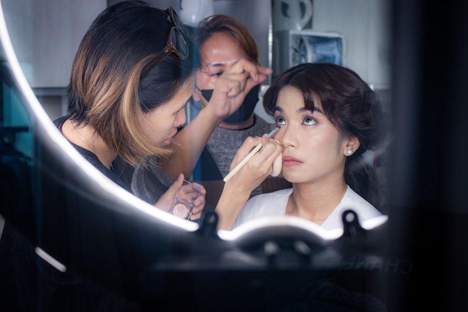 Imagem de uma profissional de maquiagem realizando uma maquiagem para uma cliente, com atenção aos detalhes e foco na preparação estética.
