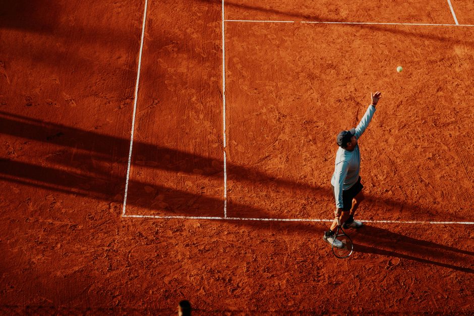 Pessoa jogando tênis em uma quadra de saibro ao entardecer, com sombra longa e movimento em ação.
