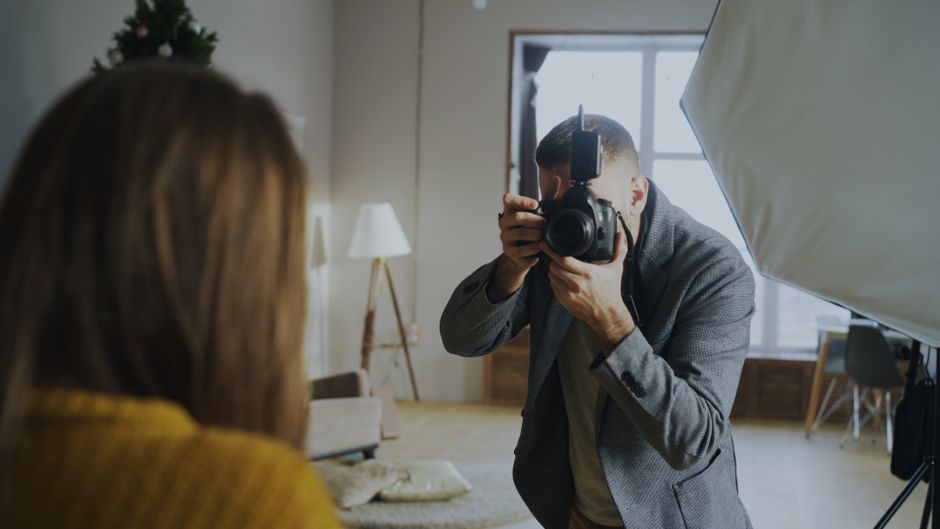 Fotógrafo profissional realizando uma sessão de fotos em um ambiente interno bem iluminado, com foco na captura de uma imagem de uma pessoa com cabelo castanho e roupa amarela.
