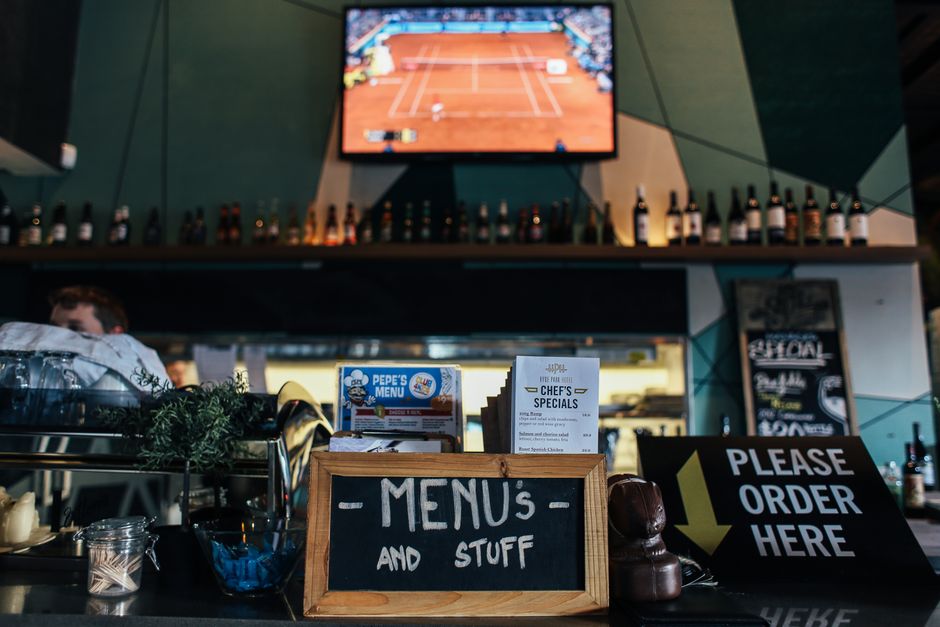 Imagem de um balcão de restaurante com um quadro de menu manual e uma placa pedindo para fazer pedidos, com destaque para um monitor de TV ao fundo exibindo uma partida de tênis.