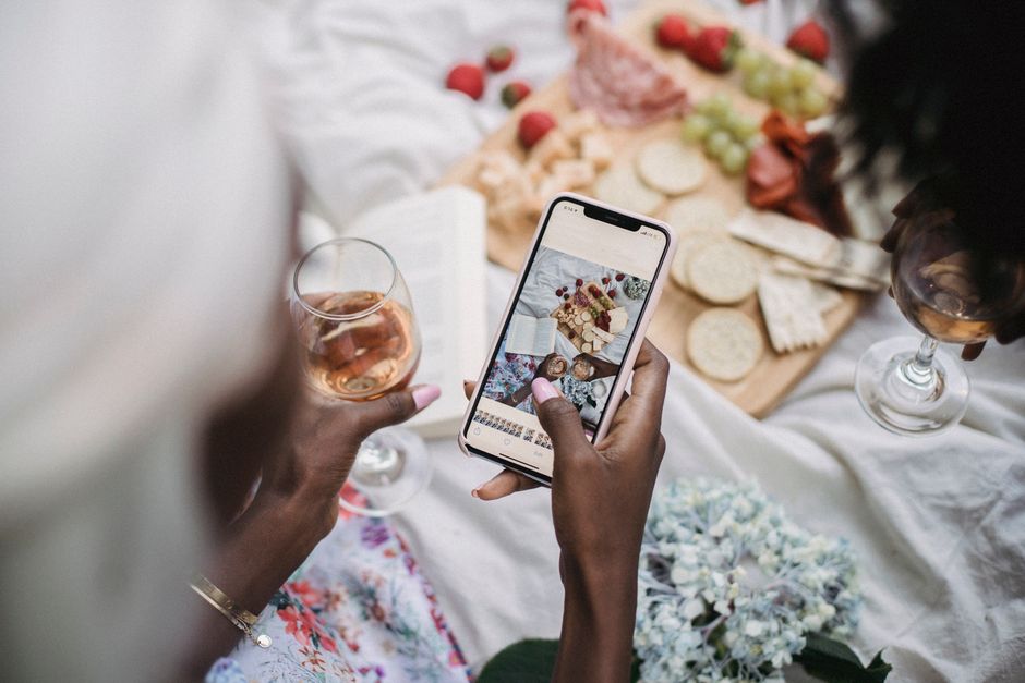 Pessoa tirando uma foto de uma mesa com comida e bebidas durante um momento social descontraído para postar em um blog de celebrações ou lifestyle.