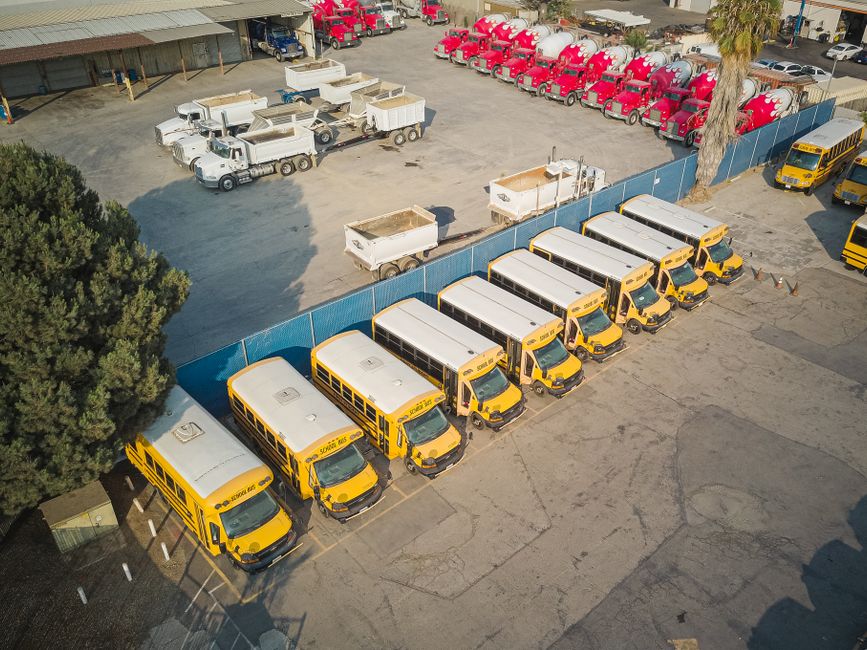 Imagem de uma frota de ônibus escolares amarelos estacionados em um pátio, com outras vans brancas e caminhões vermelhos ao fundo, indicando um ambiente de transporte escolar ou manutenção de veículos.
