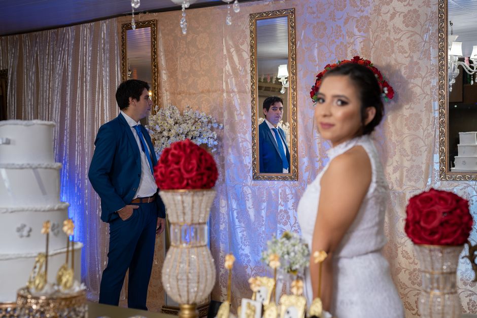 Casal de noivos em um casamento, com o noivo refletido em um espelho ao fundo e a noiva em foco em primeiro plano, decorado com flores e detalhes elegantes.