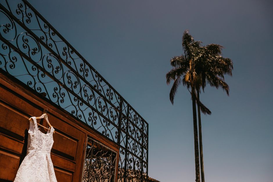 Vestido de noiva pendurado em uma parede de madeira com detalhes artísticos, sob um céu claro ao lado de uma palmeira, indicando um cenário de casamento ao ar livre.