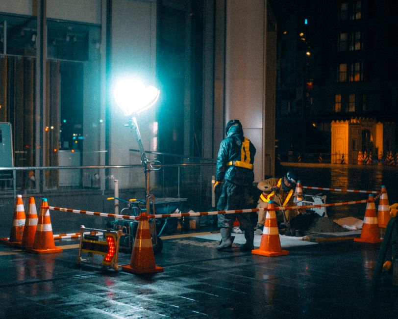 Profissionais trabalhando em uma construção noturna com iluminação artificial em uma cidade, destacando o cenário de obras urbanas.