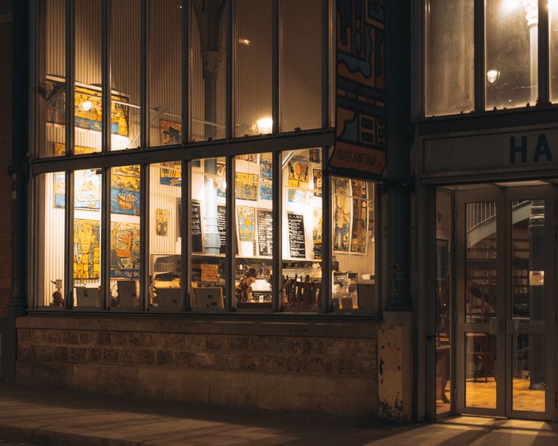 Visão externa de uma cafeteria acolhedora à noite, com iluminação quente e decoração moderna, ideal para artigos sobre cultura de cafés ou experiências gastronômicas urbanas.