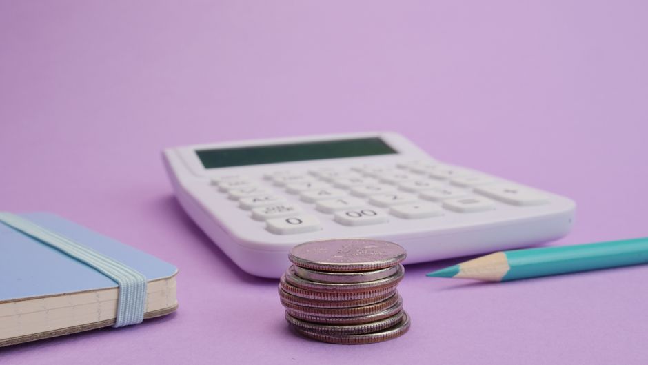Imagem ilustrativa de planejamento financeiro com calculadora, moedas, caderno e lápis sobre fundo lilás.