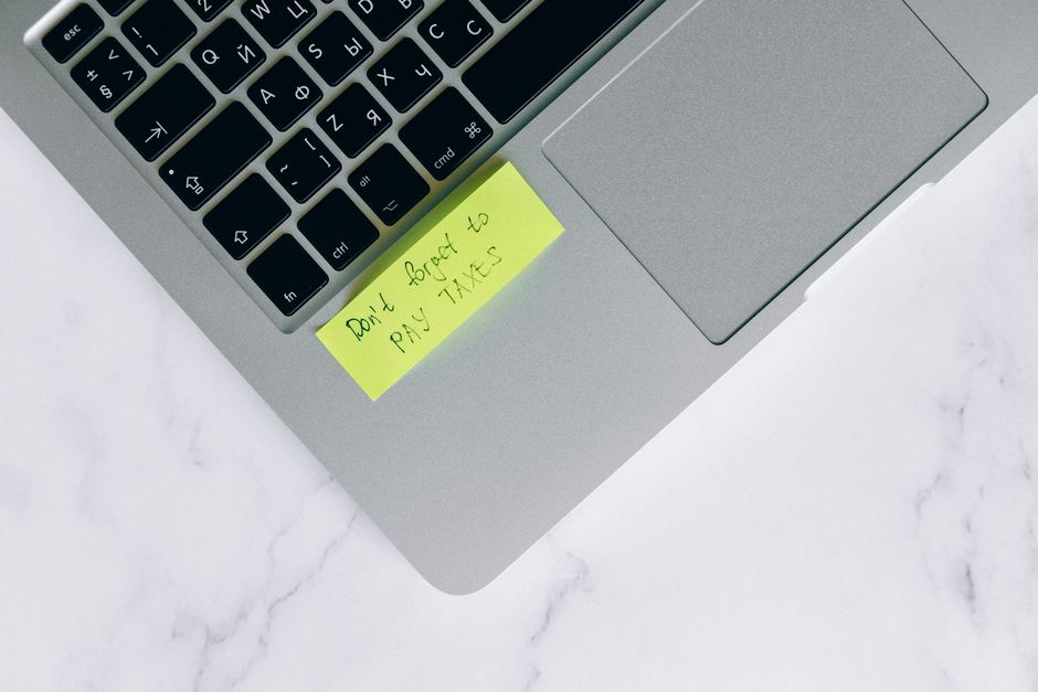 Imagem de um laptop com um bilhete amarelo dizendo 'Don't forget to pay taxes', ilustrando a importância de lembrar de obrigações fiscais.