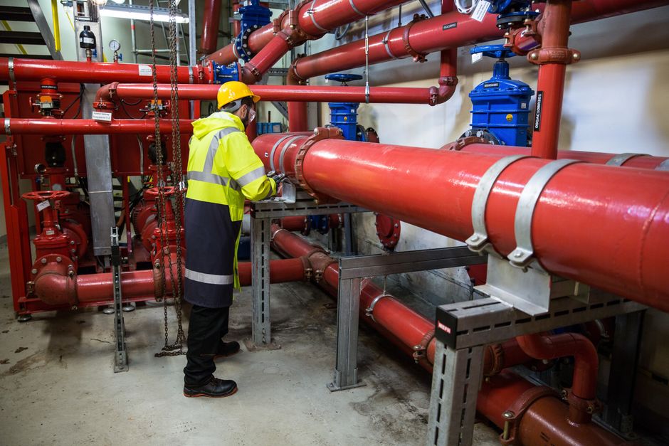 Técnico verificando ou monitorando sistemas de encanamento industrial com tubos de cores vibrantes em uma instalação mecânica.