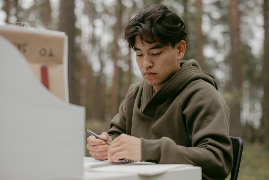 Jovem estudando ou escrevendo em um ambiente ao ar livre com árvores ao fundo, focado em suas tarefas.