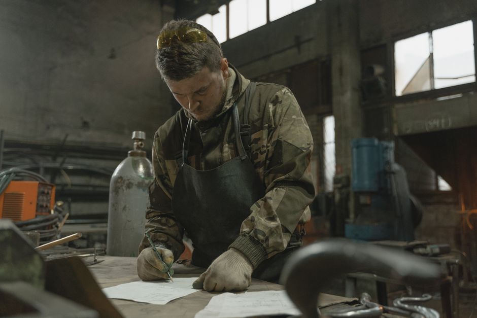 Homem usando uniforme de combate e máscara de proteção escrevendo em uma mesa em um ambiente industrial, representando resistência ou trabalho manual relacionado a atividades militares ou industriais.