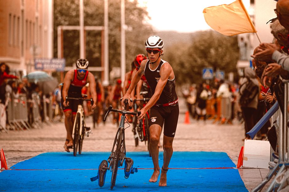 Atleta participando de uma prova de triathlon em um percurso externo, envolvendo corrida e ciclismo, com público apoiando ao redor.