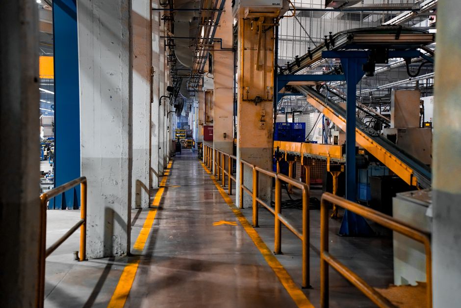 Visão de uma passarela em uma fábrica ou armazém industrial, com estrutura metálica e equipamentos de grande porte ao redor.