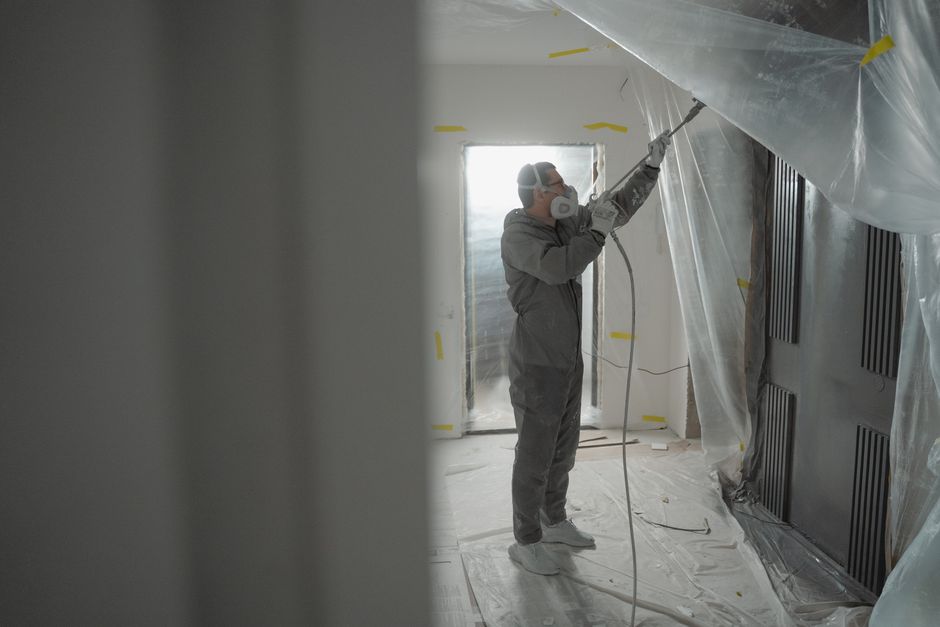 Profissional realizando pintura ou manutenção em uma parede de ambiente em reforma ou construção, com uso de máscara e equipamento de proteção.
