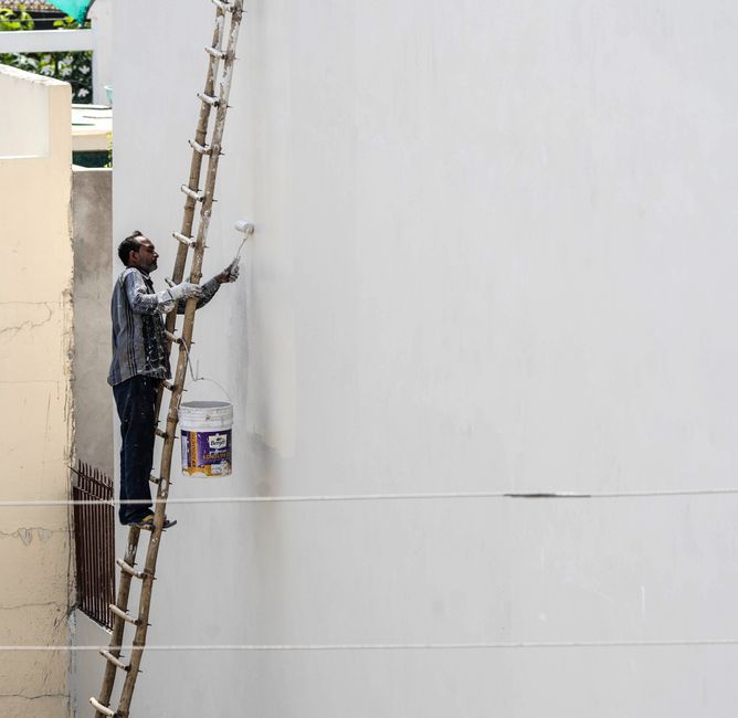 Homem pintando uma parede externa de edifício com o auxílio de uma escada, em uma atividade de reforma ou manutenção.