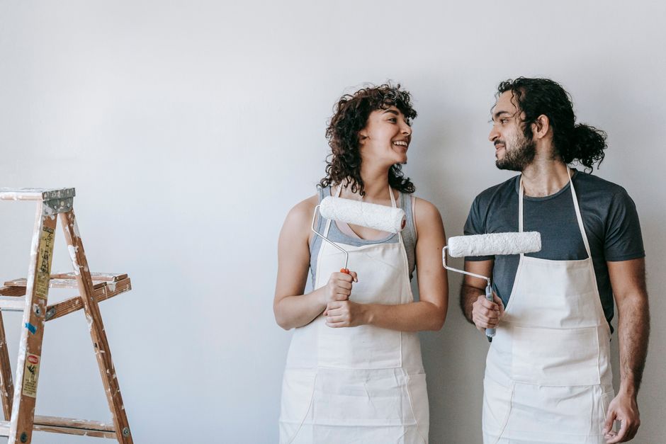 Descrição de duas pessoas sorrindo enquanto pintam uma parede, simbolizando colaboração e renovação em projeto de reforma ou decoração.