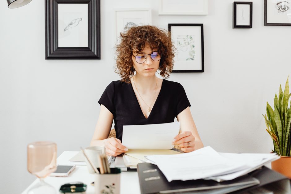 Mulher analisando documentos em um ambiente de escritório, refletindo sobre tarefas administrativas ou financeiras, com uma parede decorada ao fundo e uma planta ao lado.