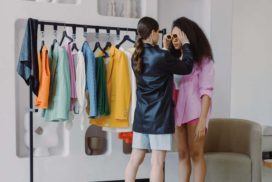 Duas mulheres discutindo sobre moda em um ambiente com roupas coloridas penduradas, sugerindo um tema de estilo e tendências.