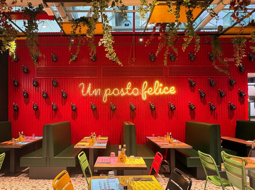 Interior de restaurante vibrante com decoração colorida, parede vermelha com lâmpadas decorativas e frase em neon 'Um posto feliz', criando um ambiente alegre e acolhedor.