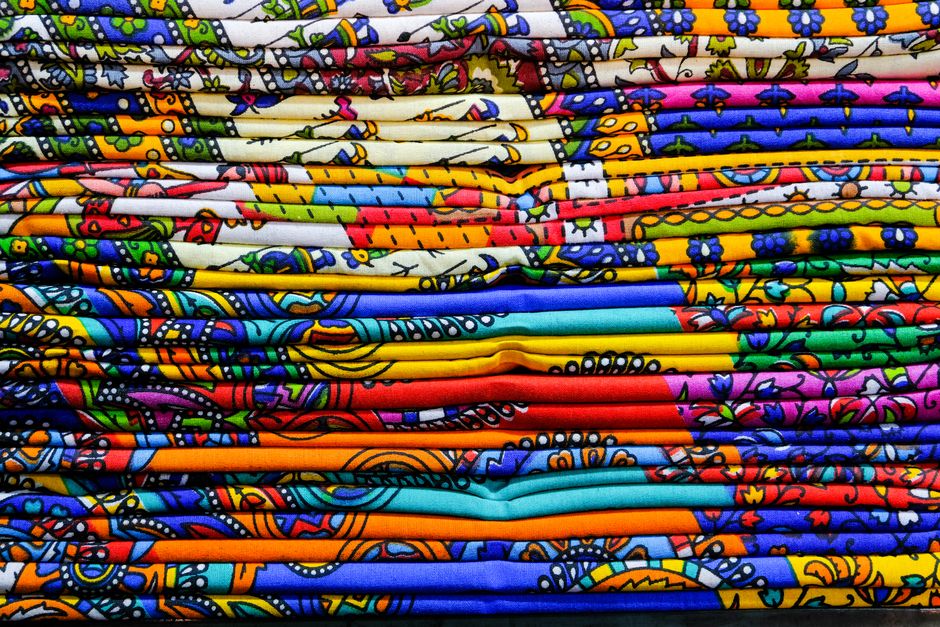 Imagem de tecidos coloridos e estampados, dispostos em camadas horizontais, prontos para confecção de roupas ou artesanato.