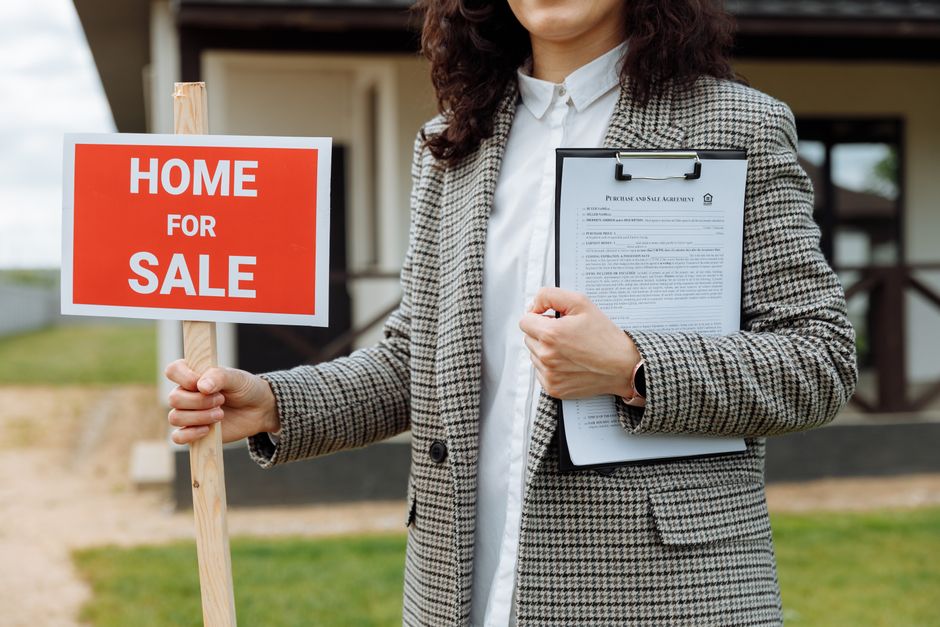 Profissional imobiliário segurando placa de venda de imóvel em frente a uma residência, representando o mercado imobiliário e negociações de compra e venda de casas.