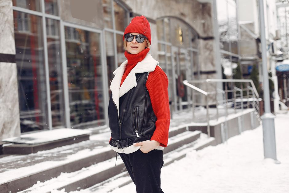 Mulher estilosa vestida com roupas de inverno, com destaque para óculos escuros e cabelo curto, caminhando na cidade durante o inverno, com decoração urbana e neve ao redor.