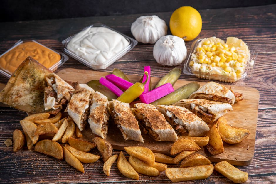 Prato de comida mexicana com burritos, batatas, picles e molhos, apresentado sobre uma tábua de madeira.