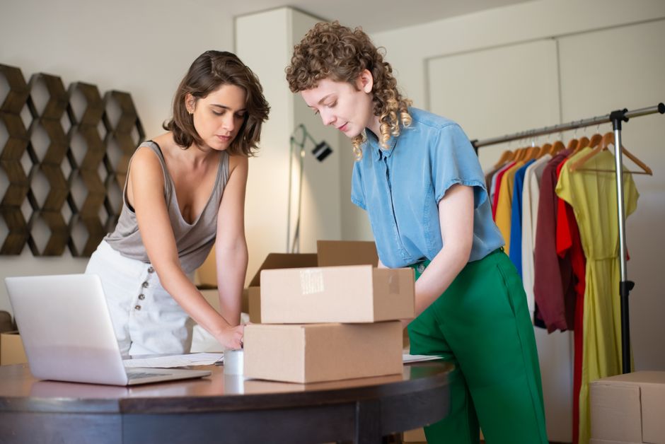 Duas mulheres analisando caixas de produtos, possivelmente em uma loja de moda ou boutique, durante uma reunião de negócios ou inventário.