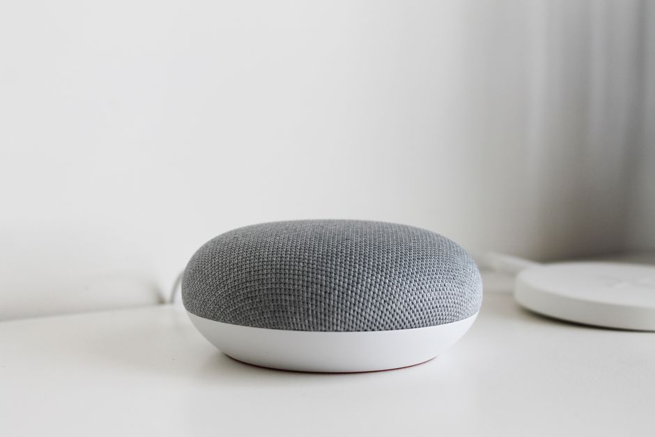 Altavoz inteligente de design moderno colocado sobre uma superfície branca, com ambiente minimalista ao fundo.