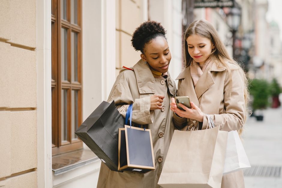 Duas mulheres fazendo compras ao ar livre, olhando algo no celular enquanto seguram diversas sacolas de lojas de roupas ou acessórios, em um ambiente urbano.