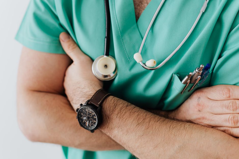 Profissional da área da saúde com braços cruzados usando uniforme médico e estetoscópio, transmitindo autoridade e cuidado em contexto hospitalar ou clínico.