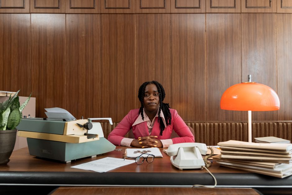 Mulher sentada em uma mesa de escritório, rodeada de documentos e equipamentos de trabalho, transmitindo concentração e profissionalismo.