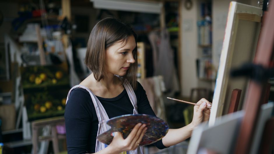 Mulher pintando em um ateliê de arte, concentrada em sua obra enquanto trabalha em uma tela em ambiente criativo.