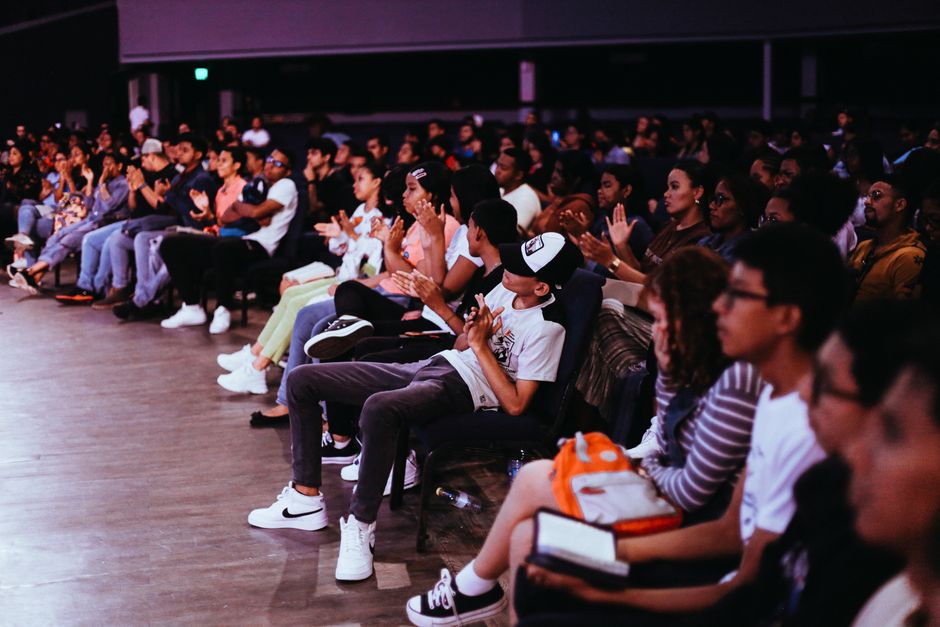 Público diversificado assistindo atentamente a uma apresentação ou palestra em um ambiente de evento indoor.