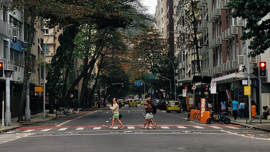 Cena urbana de uma rua movimentada com pessoas atravessando, cercada por edifícios residenciais e comerciais, com árvores e sinais de trânsito.