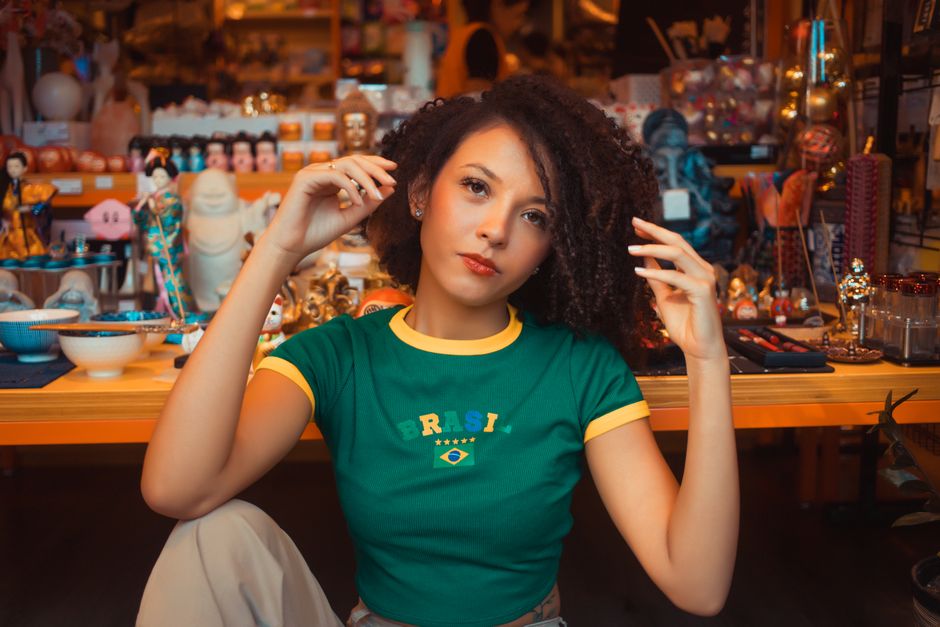 Mulher com cabelo afro e expressão confiante em ambiente de loja de souvenirs ou artigos artesanais, com destaque para sua camiseta do Brasil.