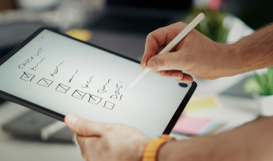 Pessoa planejando tarefas e metas utilizando um tablet com uma caneta stylus, em um ambiente de trabalho organizado.