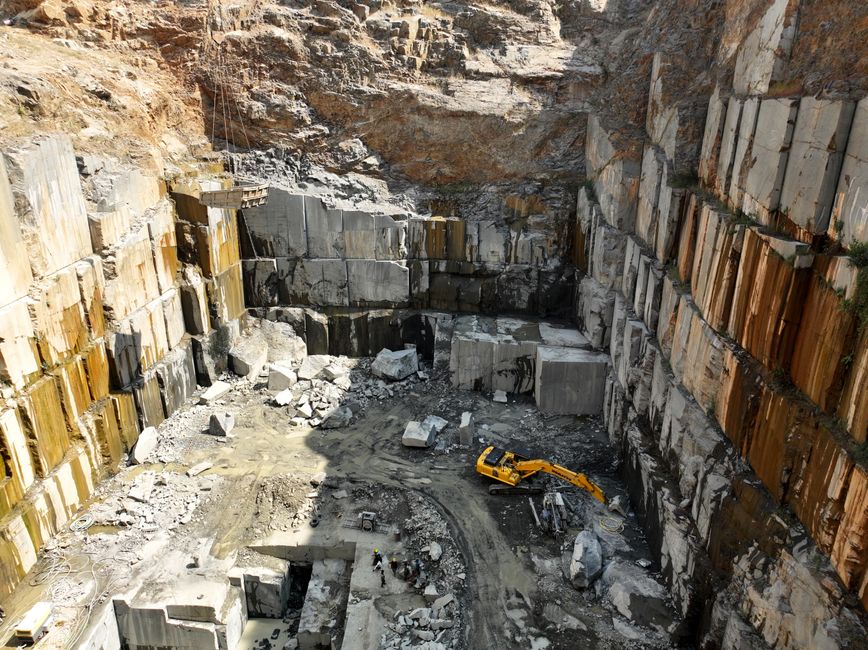 Imagem de uma grande cava de mineração, com uma escavadeira operando na etapa de extração de rochas em uma escarpa de pedra.