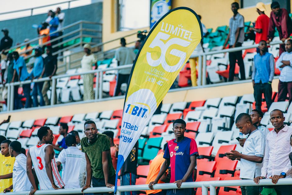 Jovens em estádio assistindo a evento esportivo, com destaque para uma bandeira publicitária de tecnologia móvel 5G.