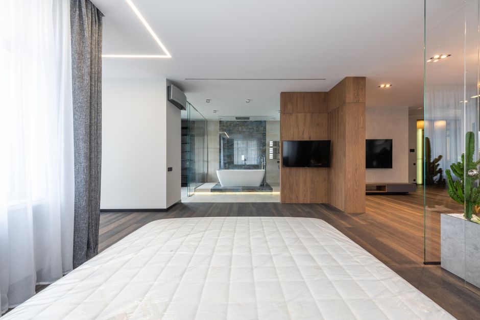 Imagem de um quarto moderno com decoração minimalista, papel de parede de cimento, janelas com cortinas e uma sala ao fundo com banheira e televisão.