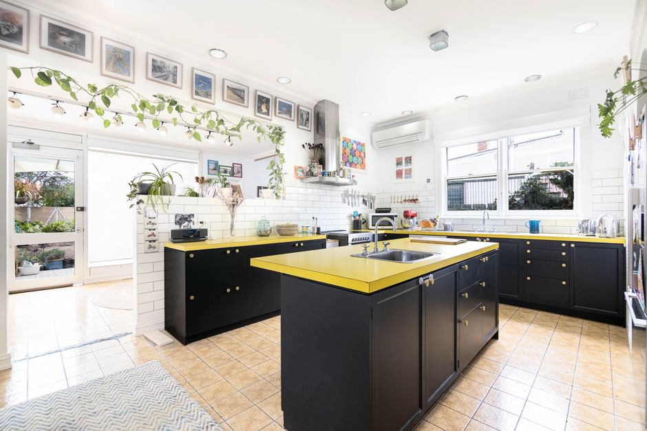 Cozinha moderna com chão de azulejo, armários pretos com detalhes amarelos, janelas grandes e decoração com plantas e fotos na parede.