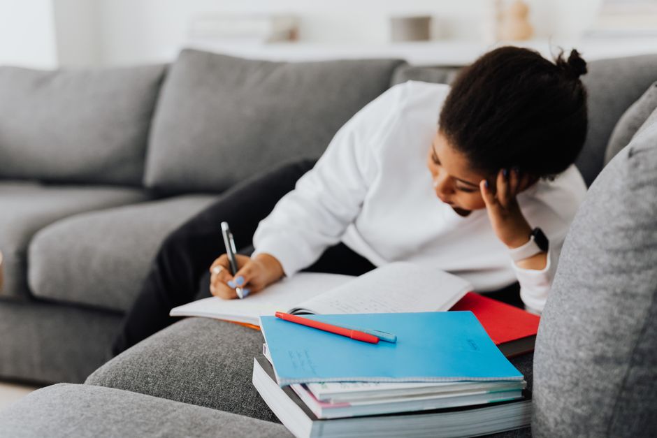 Mulher estudando e fazendo anotações em um ambiente informal, com livros e cadernos ao seu redor, em uma atmosfera de concentração e aprendizado.