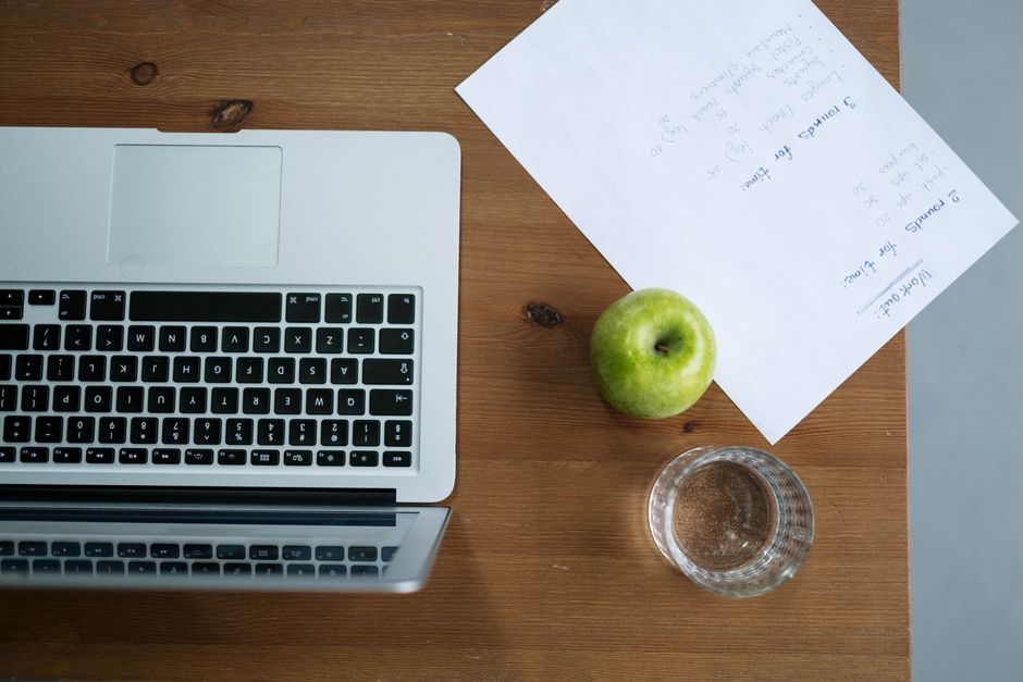 Imagem de uma mesa de trabalho com notebook, uma maçã verde, um copo de água e uma carta, ideal para ilustrar organização e planejamento de rotina no ambiente profissional ou acadêmico.