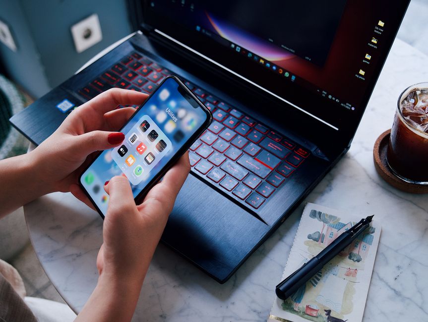Pessoa usando smartphone próxima a um laptop em um ambiente de trabalho, com itens de escritório ao redor.